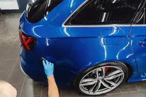 A person wearing a blue glove applies polish or ceramic coating to the rear side panel of a shiny blue car inside a garage—showcasing the quality of mobile car detailing Hillsborough County, FL residents can trust.